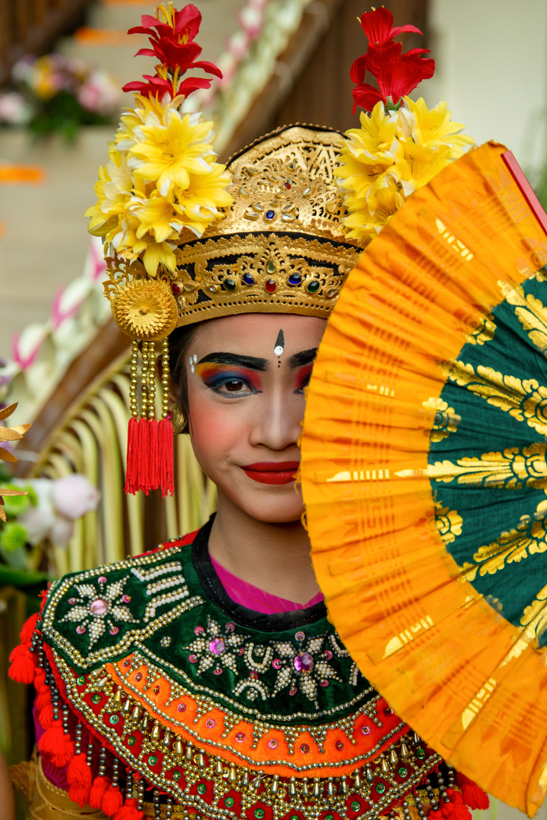 Balinese Cultural Dinner Balinese dancer dresses in traditional colorful clothes. The dancer is holding a yellow and green fan that is partly covering her face.