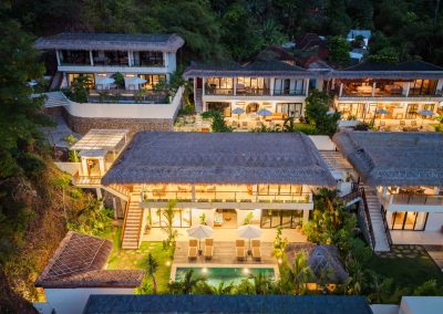 Aerial dusk view of Vesica Villas Resort in Amed, with lit thatched villas, private pools, wooden decks and white umbrellas on a forested hillside