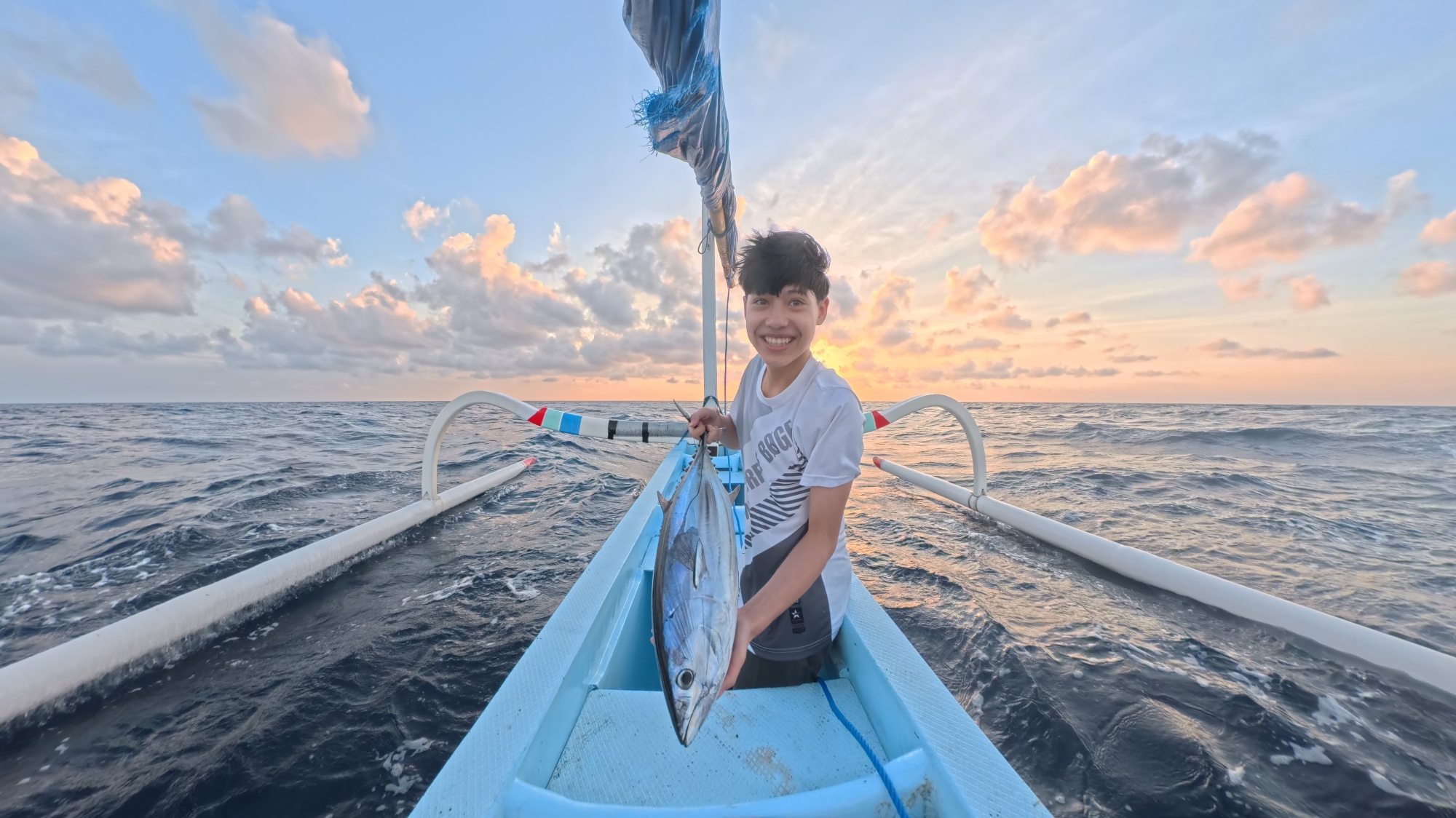 Smiling young guest holding a freshly caught tuna in a traditional jukung outrigger boat at sunrise off Amed, near Vesica Villas Resort.
