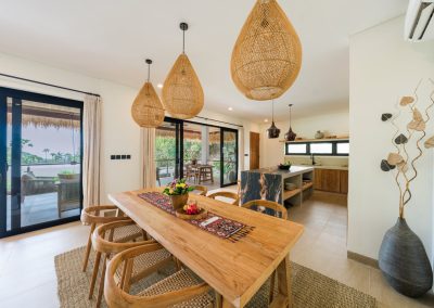 Dining area at Angin Laut at Vesica Villas Resort in Amed, with light-wood table, woven rattan pendant lights, kitchen island beyond, and open doors to a thatched outdoor terrace