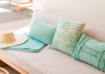 Close-up of a daybed at Angin Laut at Vesica Villas Resort in Amed, with teal pillows, cowrie-shell trim, a straw hat, and folded turquoise throw