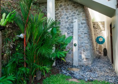 Outdoor shower on a stone wall with chrome rain head, river pebbles, and tropical plants at Angin Laut at Vesica Villas Resort in Amed