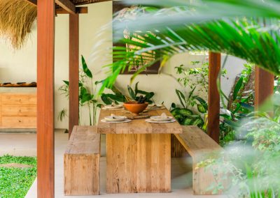 Thatched outdoor dining at Angin Laut at Vesica Villas Resort in Amed, with a set wooden table and benches, tropical plants, and cream wall art beyond