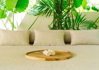Daybed at Angin Laut at Vesica Villas Resort in Amed, with beige cushions, wood tray of rolled towels, and tropical greenery behind a low wall