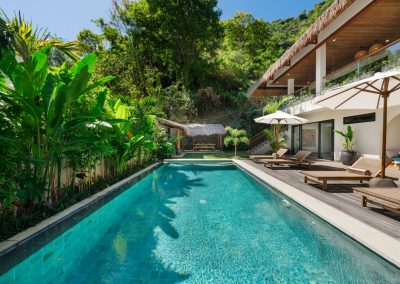 High-angle view of the turquoise pool at Angin Laut at Vesica Villas Resort in Amed, with timber deck loungers, white umbrellas, thatched roof, and forested hillside