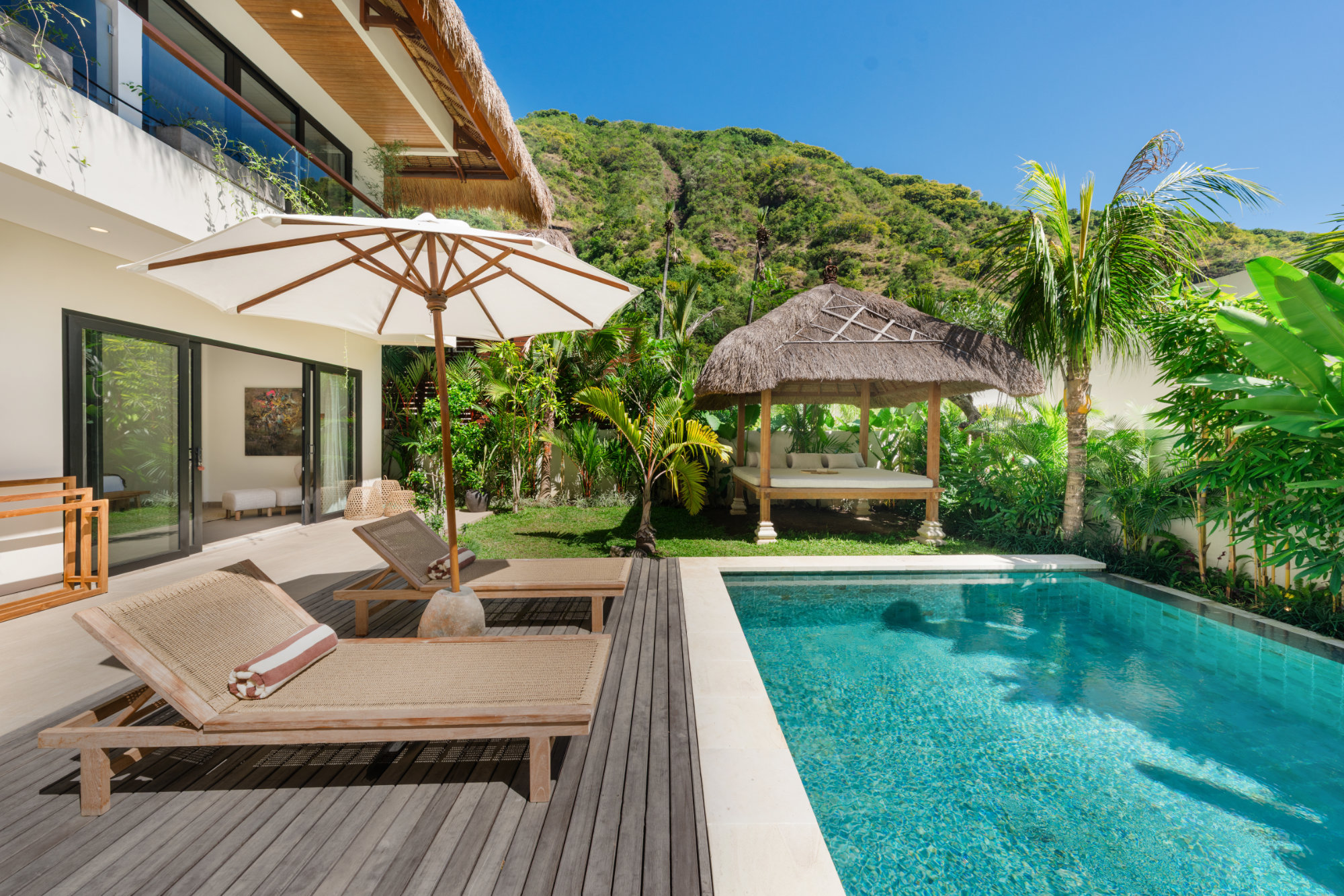 Angin Laut Pool Sun-beds with umbrella facing a turquoise pool in a luxury residence at the Vesica Villas Resort in Amed. A traditional bale day-bed with alang-alang roof sites at the end of the pool and tropical hills are visible in the background.