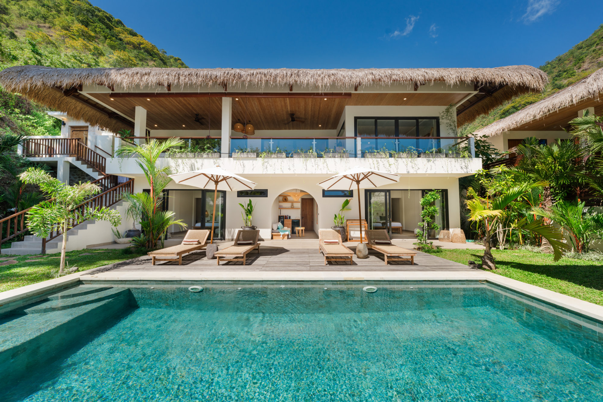 Angin Laut Pool Luxury resort villa in Amed built over two floors. A large turquoise swimming pool with wooden sunbeds surrounded by a tropical garden. The sky behind the villa is clear blue with tropical hills visible at both sides.
