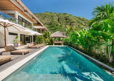 Private turquoise pool courtyard at Angin Laut villa at Vesica Villas Resort in Amed, with timber deck, loungers, white umbrellas, thatched bale gazebo, and lush hillside behind