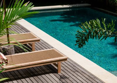 High-angle pool corner at Angin Laut at Vesica Villas Resort in Amed, with two wood loungers, striped towel, timber deck, and overhanging palm fronds