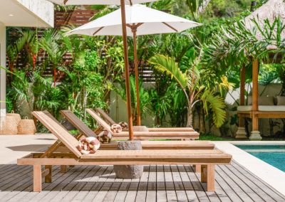 Timber deck at Angin Laut at Vesica Villas Resort in Amed, with wood loungers under white umbrellas beside the turquoise pool, thatched bale, and tropical foliage