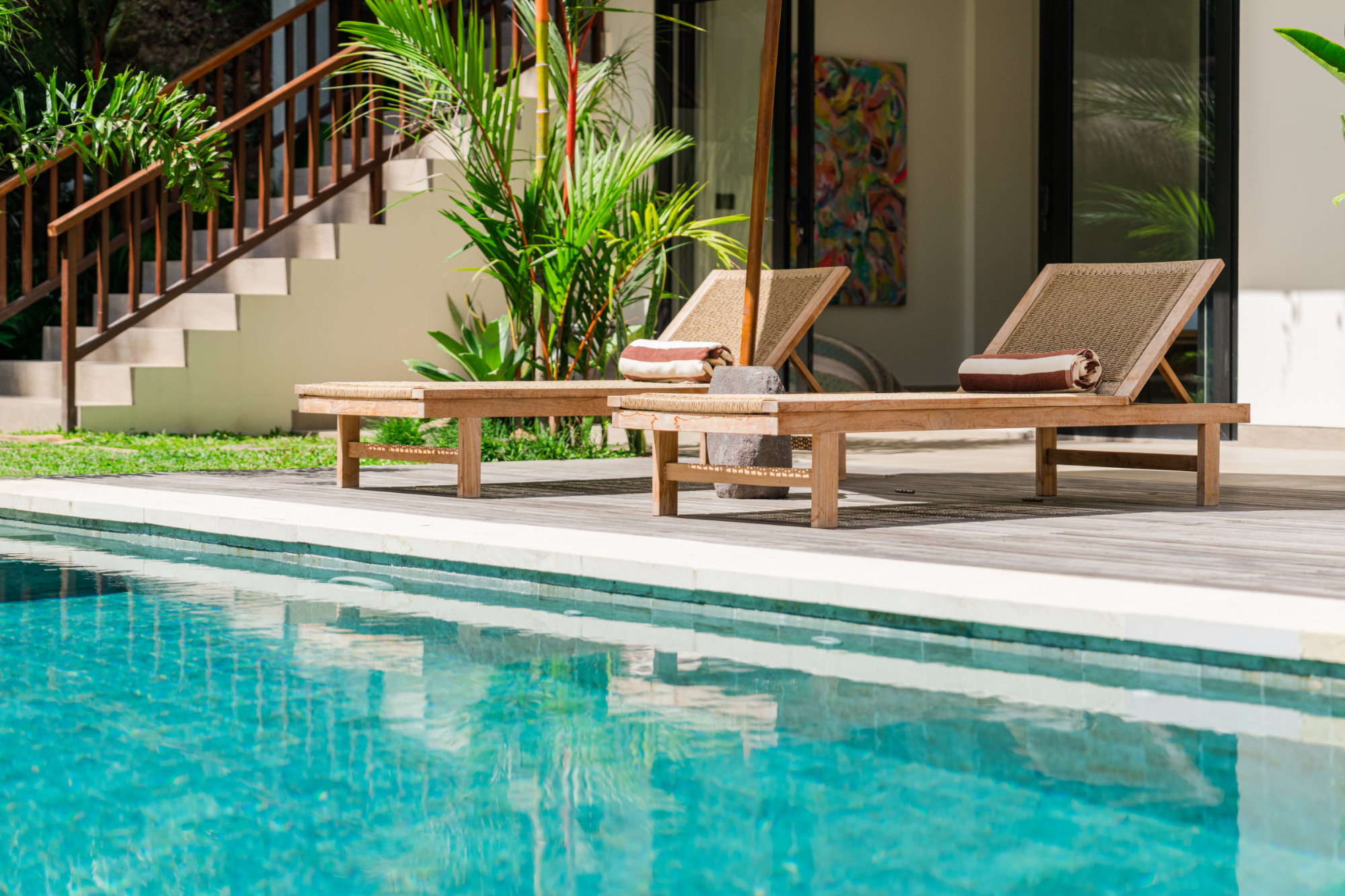 Poolside loungers with striped towels on a timber deck at Angin Laut at Vesica Villas Resort in Amed, beside black-framed glass doors and white stairs