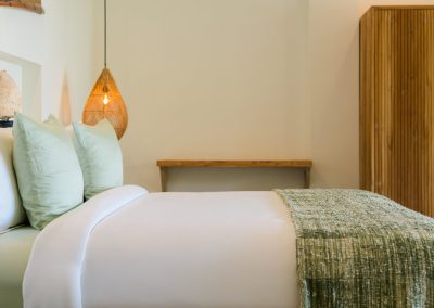 Close-up of Angin Laut bedroom linens at Vesica Villas Resort in Amed, white sheets with sage pillows, woven olive throw, rattan pendant lamp, and tall slatted wood wardrobe