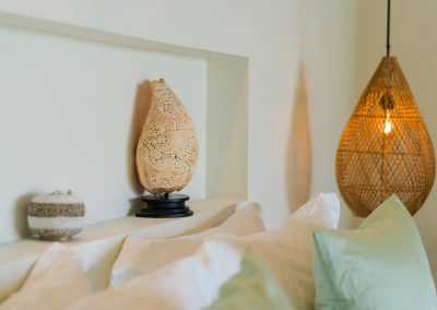 Bedroom wall niche at Angin Laut at Vesica Villas Resort in Amed, with carved shell decor and striped pot above sage pillows beside a rattan pendant