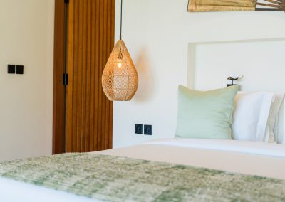 Bedroom linens at Angin Laut at Vesica Villas Resort in Amed, white sheets with sage pillow and olive throw beside a lit rattan pendant and slatted wood panel