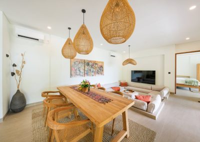Bright living and dining room at Angin Laut at Vesica Villas Resort in Amed, with wooden table under rattan pendants, L-shaped sofa, abstract wall art, and a doorway to a bedroom