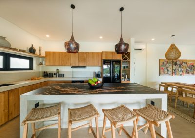Kitchen island at Angin Laut at Vesica Villas Resort in Amed, with dark marbled countertop, fruit bowl, four woven bar stools, copper pendants, and dining table beyond