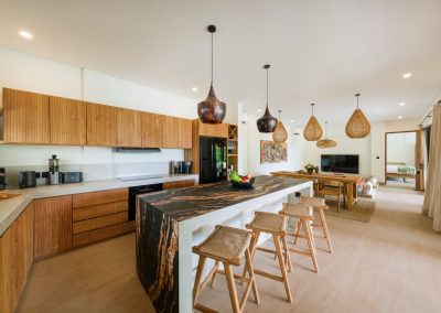 Open-plan kitchen and dining at Angin Laut at Vesica Villas Resort in Amed, with marbled island, woven bar stools, wood dining table under rattan pendants, and living area beyond