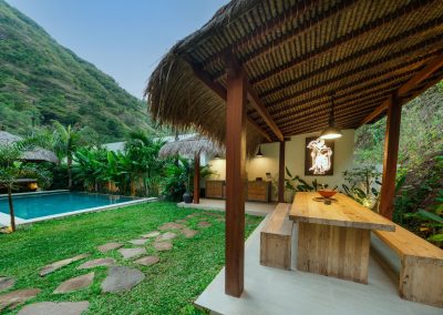 Thatched outdoor dining pavilion at Angin Laut at Vesica Villas Resort in Amed, with wooden table and benches, backlit wayang art, pool, lawn stepping stones, and hillside