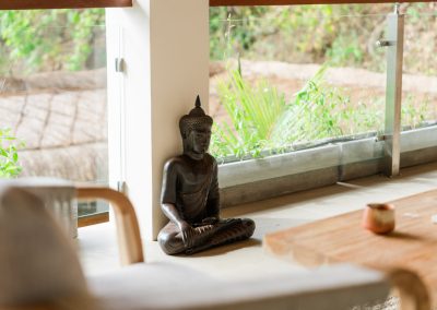 Dark seated Buddha statue on the tiled floor at Angin Laut at Vesica Villas Resort in Amed, beside a white pillar and glass doors opening to tropical greenery
