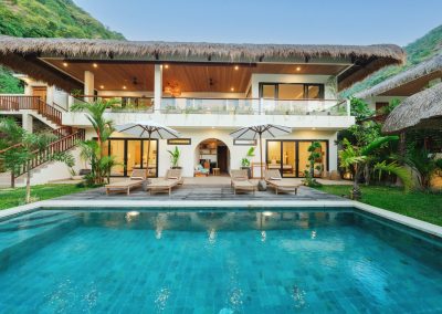 Two-story thatched Angin Laut villa at Vesica Villas Resort in Amed, at dusk, with pool, wooden loungers, white umbrellas, and a warm-lit open upper terrace