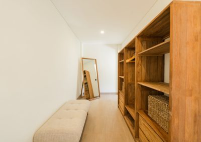 Dressing closet at Angin Laut at Vesica Villas Resort in Amed, with a light-wood open wardrobe, cream tufted bench, and a full-length wooden-framed mirror