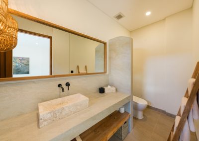 Bathroom at Angin Laut at Vesica Villas Resort in Amed, with a stone vessel sink vanity, wood-framed mirror, toilet behind a partition, and a wooden towel ladder