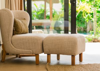 Beige armchair and ottoman with olive cushion beside a black-framed glass door to the garden at Angin Laut at Vesica Villas Resort in Amed