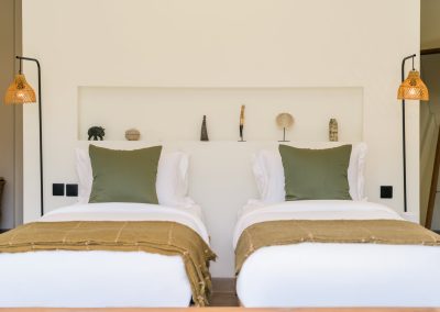 Twin beds with white linens, olive pillows, and mustard runners flanked by woven rattan floor lamps at Angin Laut at Vesica Villas Resort in Amed
