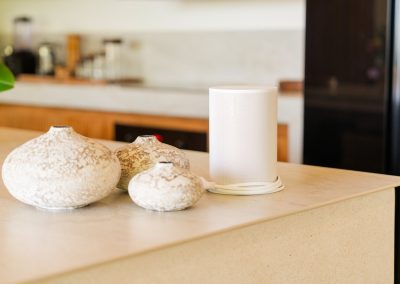 Kitchen counter vignette at Aman Amed at Vesica Villas Resort in Amed, with three speckled ceramic bud vases beside a white smart speaker, light wood cabinets and a black refrigerator softly blurred behind