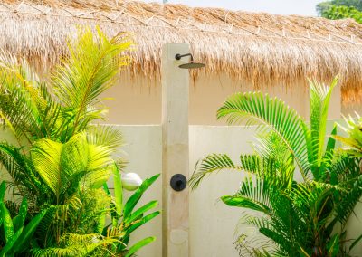 Outdoor rain shower at Aman Amed at Vesica Villas Resort in Amed, with a cream stone pillar, matte black rain head and control, tropical planting on both sides, white wall, and thatched roof edge above