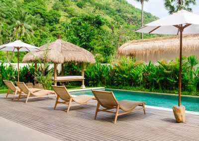 High-angle view of the private pool at Aman Amed at Vesica Villas Resort in Amed, with four woven lounge chairs and twin white umbrellas on a wood deck, a thatched bale daybed across turquoise water, and a steep jungle hillside beyond