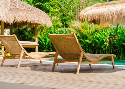 Two woven lounge chairs on a light wood deck beside the turquoise pool at Aman Amed at Vesica Villas Resort in Amed, with a thatched-roof bale, tropical plants, and orange-red flowers in bright daylight