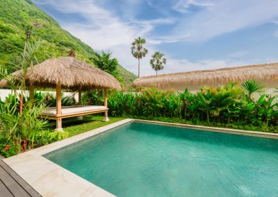 Thatched Balinese bale with white daybed beside the rectangular turquoise pool at Aman Amed villa at Vesica Villas Resort in Amed, framed by tropical planting and a steep green mountain under a blue sky