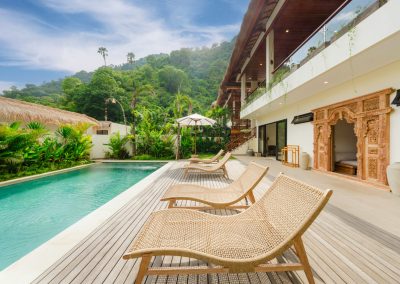 Pool deck with woven loungers and a white umbrella at Aman Amed at Vesica Villas Resort in Amed, facing the two-story white villa with carved Balinese door, glass doors, upper balcony, and forested hillside backdrop