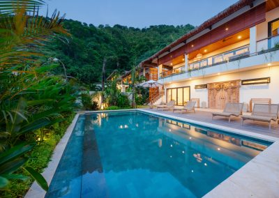 Dusk view of the private pool and wooden deck at Aman Amed at Vesica Villas Resort in Amed, with warm villa lights, carved ground-floor door, upper balcony lamps, lounge chairs, and a lush mountain rising behind