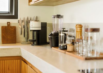 Kitchen counter corner at Aman Amed at Vesica Villas Resort in Amed, showing a magnetic knife strip, cutting board, coffee machine, blender, French press tray, and glass canisters on stone above ribbed wood cabinets