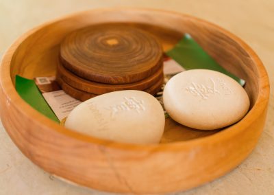 Wooden welcome bowl at Aman Amed at Vesica Villas Resort in Amed, holding engraved white stones reading Honesty and Love, stacked dark wood coasters, and printed guest cards on a stone surface
