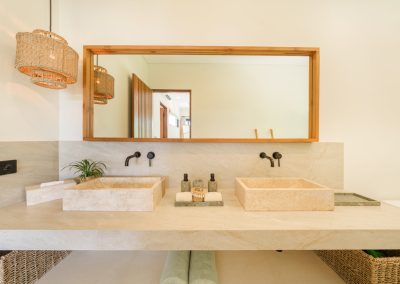 Double stone vessel sinks on a floating vanity at Aman Amed at Vesica Villas Resort in Amed, with matte black wall faucets, a wide wood-framed mirror, woven pendant light, toiletry trays, and rolled towels below
