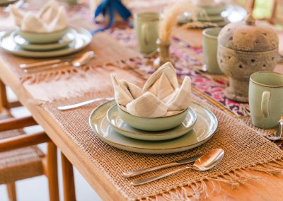 Close-up place setting at Aman Amed villa at Vesica Villas Resort in Amed, with sage green ceramic plates and bowl, a checkered napkin crown, silver cutlery, and a patterned woven runner on the wood table