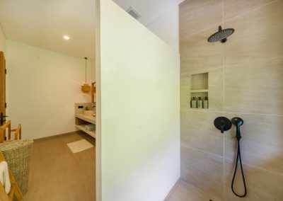 Walk-in shower behind frosted glass at Aman Amed at Vesica Villas Resort in Amed, with matte black rain head and handheld, beige stone tiling, toiletry niche, and vanity with woven pendant visible beyond