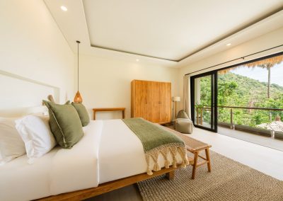 Bright bedroom at Aman Amed villa at Vesica Villas Resort in Amed, showing a wood-frame bed with olive accents, woven bench, wardrobe and desk, beanbag by the glass door, and a balcony over a lush hillside