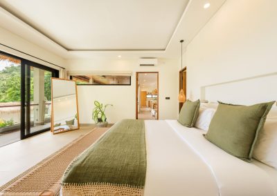 Bedroom at Aman Amed at Vesica Villas Resort in Amed, with white linens, sage pillows and runner, woven bench, full-length wood mirror by open black-framed doors, and a tropical hill view from the balcony