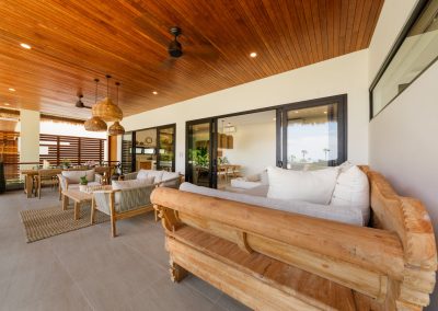 Long covered terrace at Aman Amed at Vesica Villas Resort in Amed, from a carved daybed past a rope-detail lounge set to a dining table under woven pendants, with black-framed doors into the bright interior