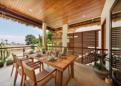 Outdoor dining terrace at Aman Amed at Vesica Villas Resort in Amed, with a set wood table for six, sage ceramics, pillar candles, woven chairs, thatched roof edge, wood-slat privacy screen, and glass railing over trees