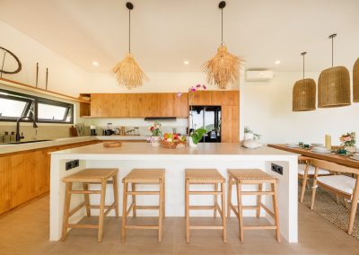 Open-plan kitchen at Aman Amed at Vesica Villas Resort in Amed, with a white island, four rattan bar stools, fruit bowl and orchid, shaggy straw pendants, ribbed wood cabinets, black fridge and hood, and dining table to the right