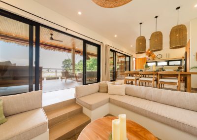 Sunken lounge and dining at Aman Amed at Vesica Villas Resort in Amed, with built-in beige seating, round wood coffee table, dining chairs under woven cylinder pendants, and open black-framed doors to a covered terrace