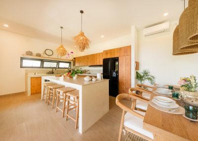 Kitchen and dining at Aman Amed at Vesica Villas Resort in Amed, with a white island and rattan stools under shaggy straw pendants, ribbed wood cabinets and black fridge, and a set wood table with woven chairs in the foreground