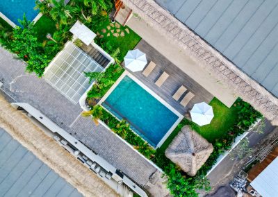 Aerial top-down view of the private turquoise pool at Aman Amed at Vesica Villas Resort in Amed, with a dark wood deck of loungers under white umbrellas, lawn, thatched bale, and tropical planting between thatched roofs