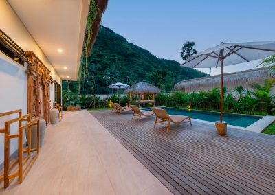 Pool courtyard at dusk at Aman Amed at Vesica Villas Resort in Amed, with wood deck loungers, white umbrella, thatched bale beyond the pool, carved wood door detail on the villa wall, and a steep forested mountain behind