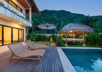 Twilight pool view at Aman Amed villa at Vesica Villas Resort in Amed, with woven loungers on the wood deck, lit thatched bale, two-story white villa with balcony vines and warm interior light, and a forested mountain beyond